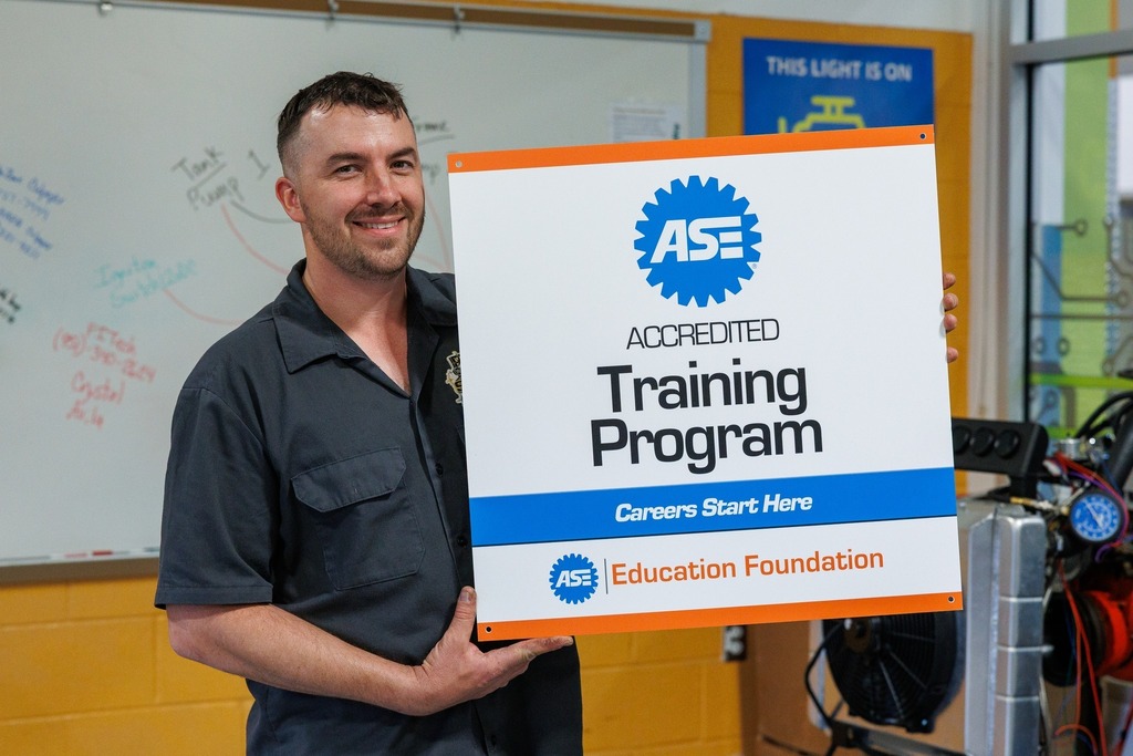 An automotive instructor stands in a classroom holding an ASE Accredited Training Program sign from the ASE Education Foundation, with automotive equipment and classroom whiteboards visible in the background.