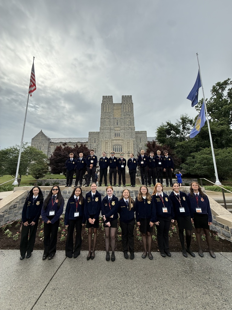 A group of students in uniform pose in front of a large building with flags and greenery in the background.