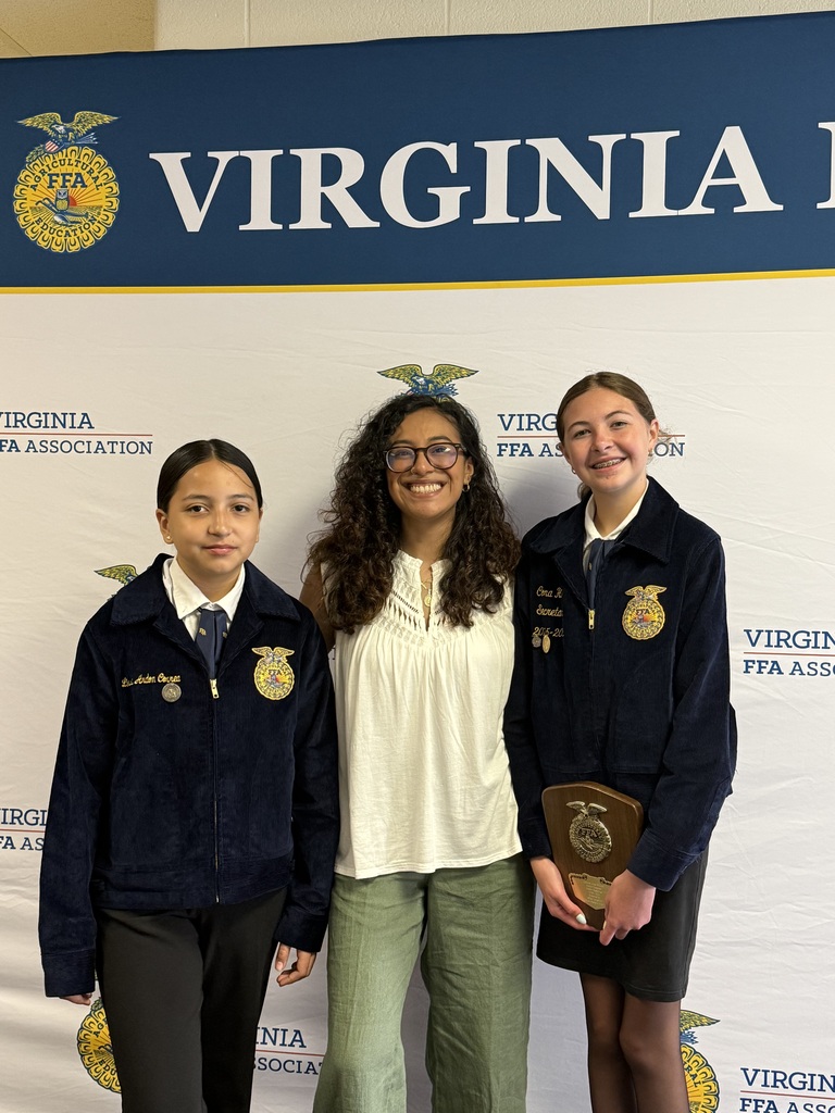 Three women stand behind a banner. Two hold a trophy, one in each hand. All wear jackets and glasses.