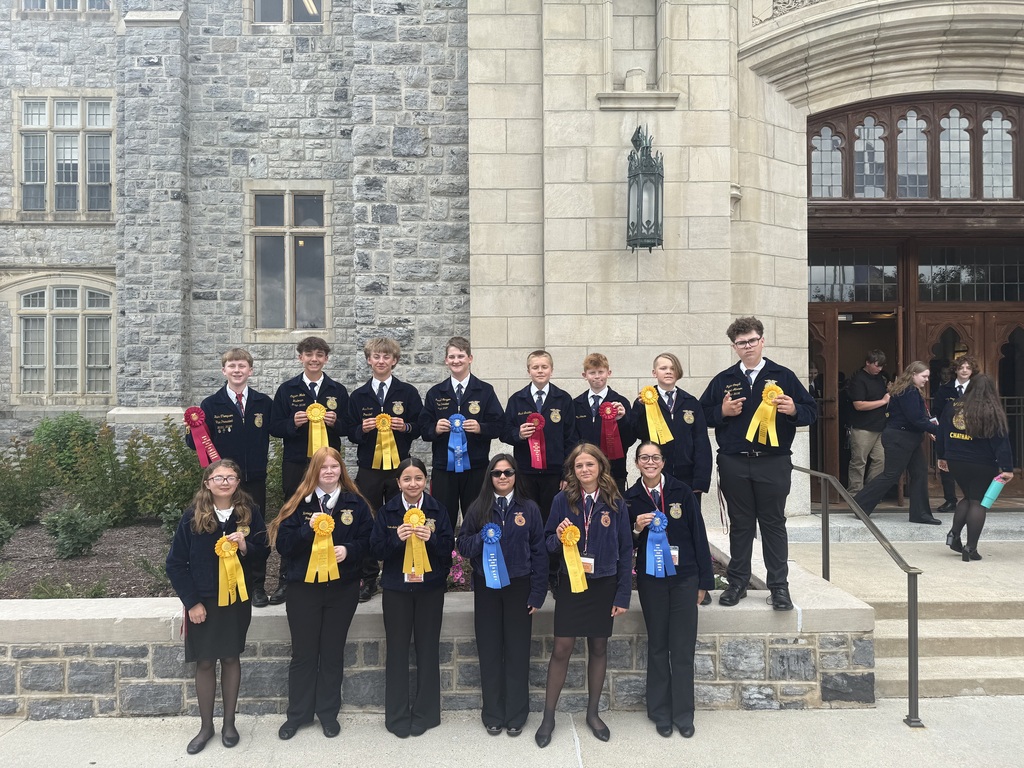 Students in formal attire with ribbons pose in front of a building with arched windows.