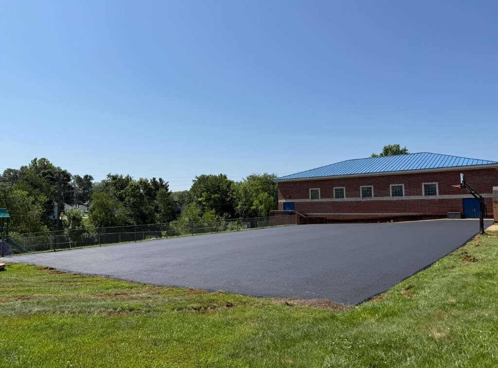 A large building with a blue roof stands on a grassy field. A paved basketball court is in front.