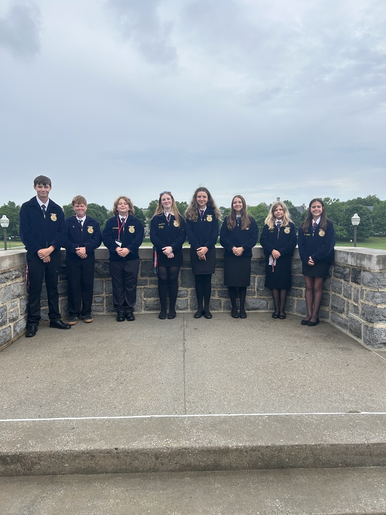 Eight Culpeper Middle School FFA members wearing official blue FFA jackets pose together on the Virginia Tech campus during the 100th Annual Virginia FFA Convention. The group stands on a stone overlook with trees and campus buildings visible in the background beneath an overcast sky.