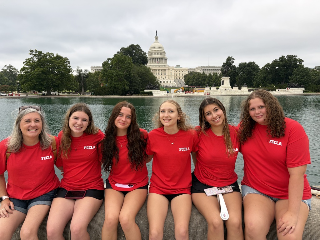 A group of seven women in matching red t-shirts, black shorts, and sandals pose near a lake with trees and a building.