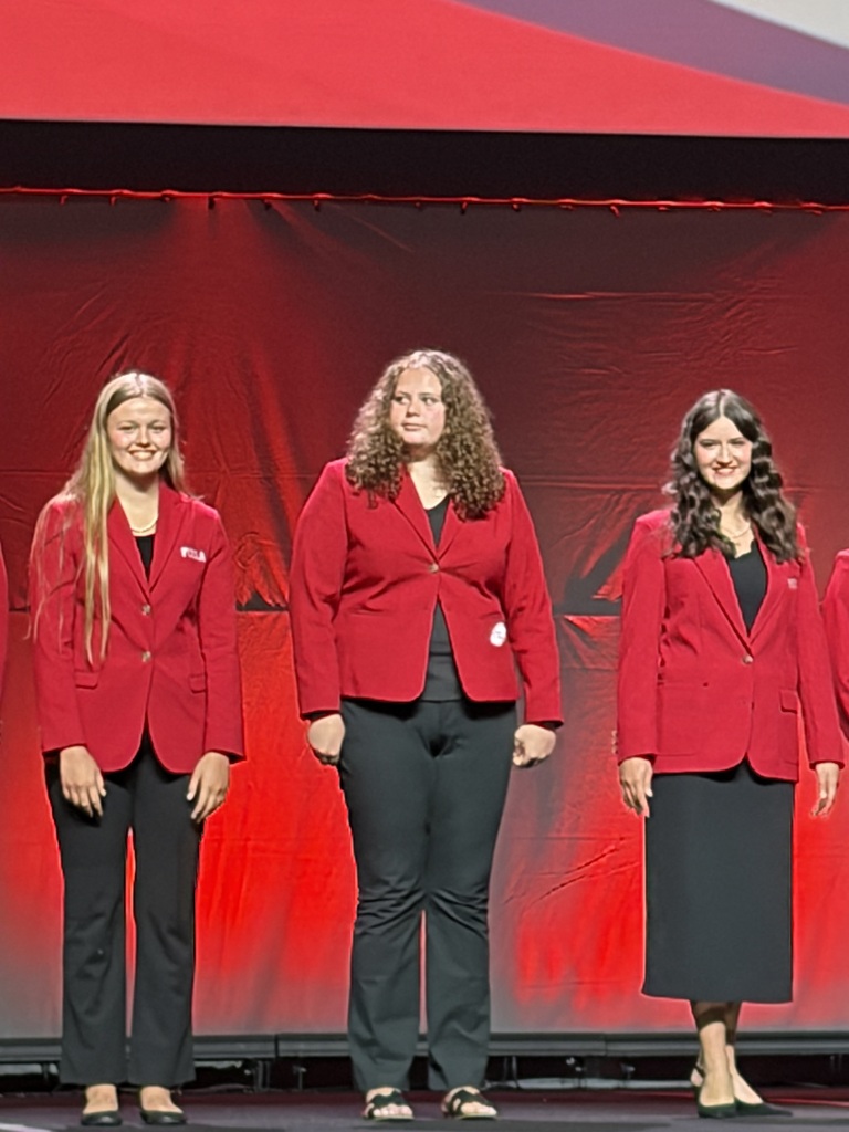 Four women wearing red blazers and black pants or skirts stand on a stage. The stage has a red backdrop.