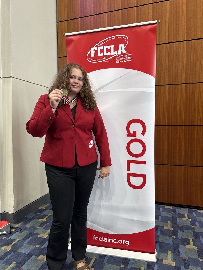 A woman stands before a red and white banner that reads "FCCLA GOLD" with "fcclainc.org" below. She holds a medal.