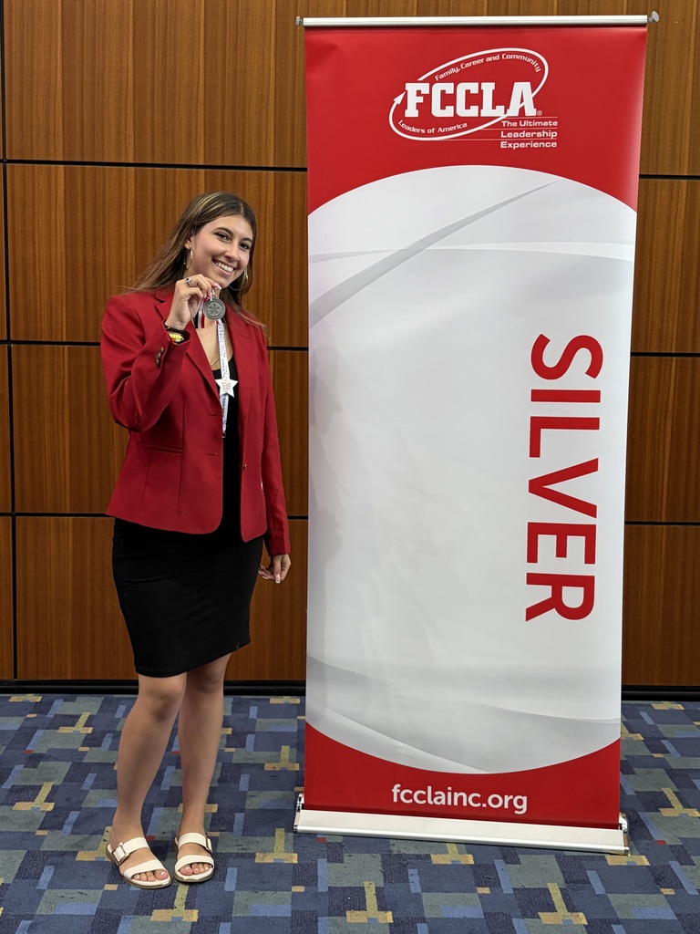 A woman in a red blazer and black skirt holds a microphone in front of a silver banner.