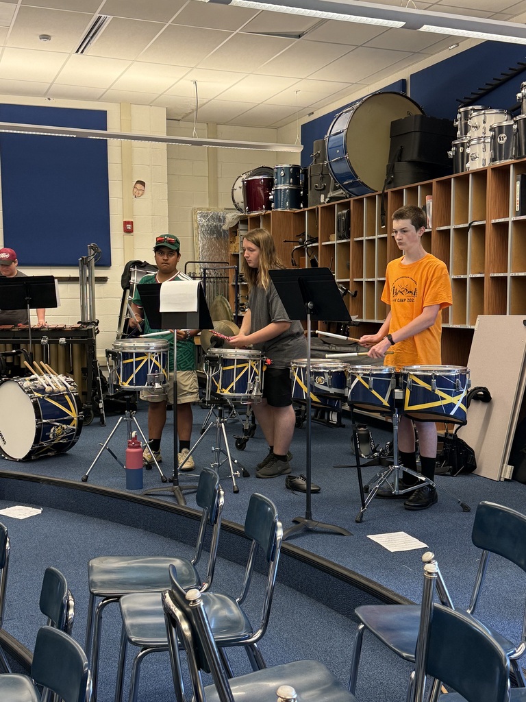Three individuals playing drums in a music room, with wooden shelves in the background.
