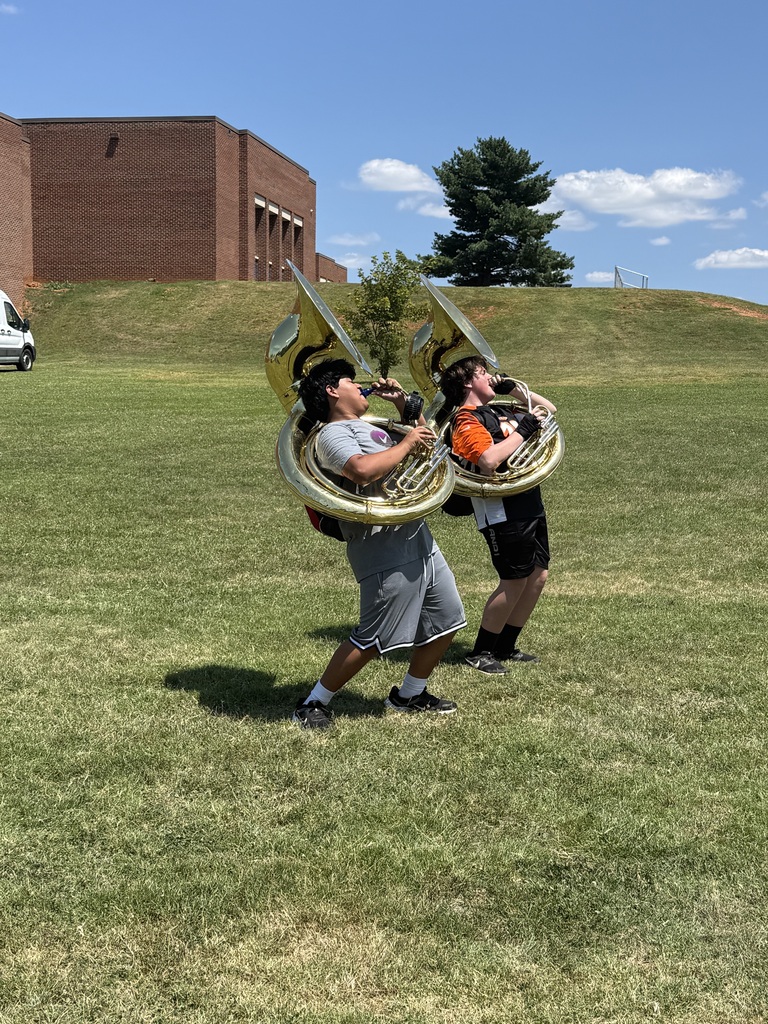 Two individuals playing brass instruments, tuba, outdoors on a grassy field. A building is in the background.