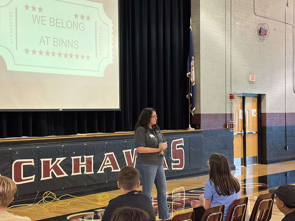 A woman in a school gym presents to a seated audience. A projector screen shows "We Belong at Binns."