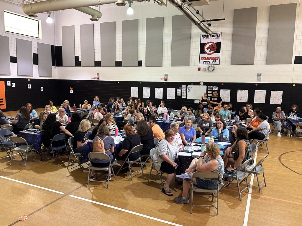 A gym with many people seated around tables, a whiteboard on the wall, and a fire extinguisher.