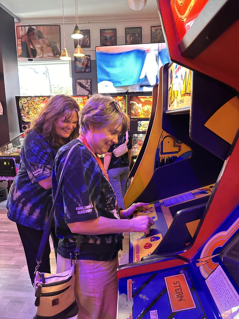 Two women stand at an arcade game with vibrant colors. One plays the game while the other watches.