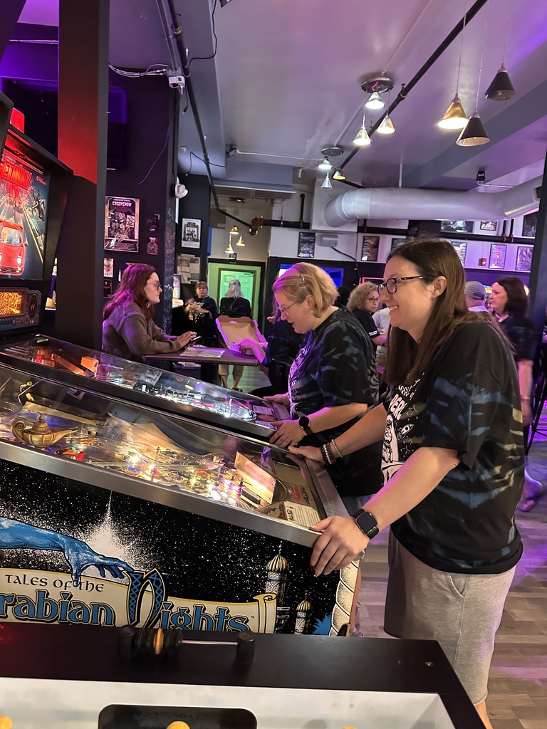 A room with pinball machines; a woman in a black shirt watches a game. Other people play behind her.