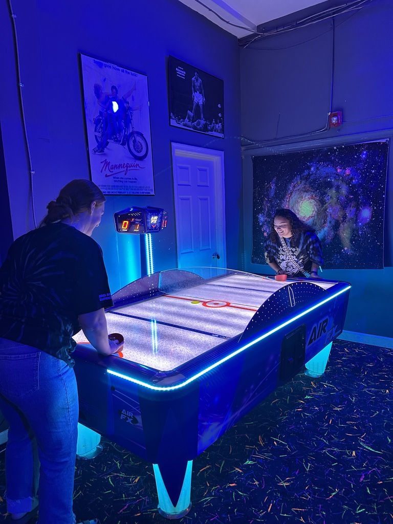 Two people playing air hockey in a room with blue lights and a large painting on the wall.