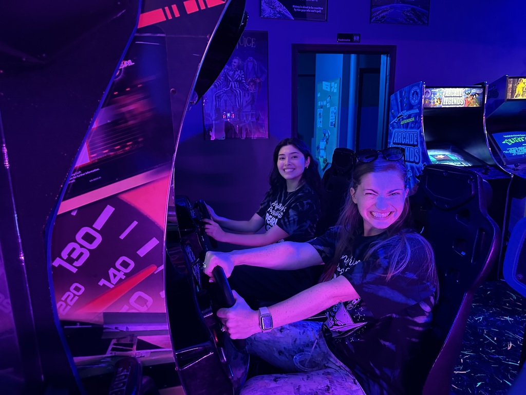 Two Pearl Sample staff members smile while seated at a racing arcade game under bright purple and blue neon lights at Throwbacks Arcade.