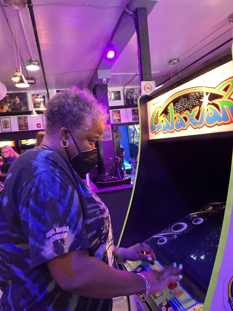 Person wearing a mask playing an arcade game in a colorful room with monitors and pictures.