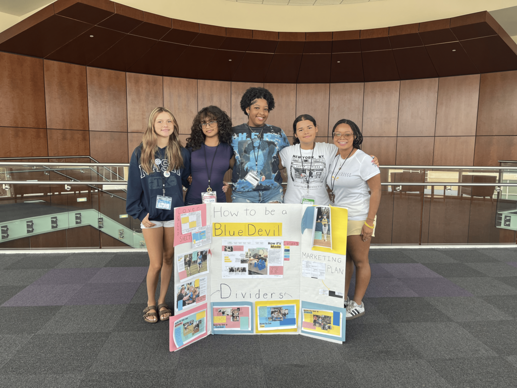 Five female students stand by a poster board with colorful pictures and text. They are inside a building with wooden walls and a metal railing.