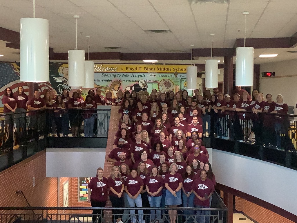 A large group of people in maroon shirts posing on a balcony, looking down at a hallway.