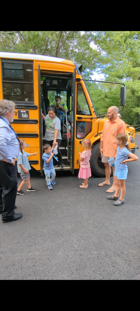 People are boarding a school bus on a sunny day. A few children are standing in front, and adults are nearby.