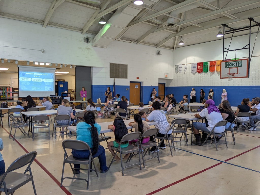 Indoor hall with tables and chairs, people seated. Screen on the wall, basketball hoop in the background.