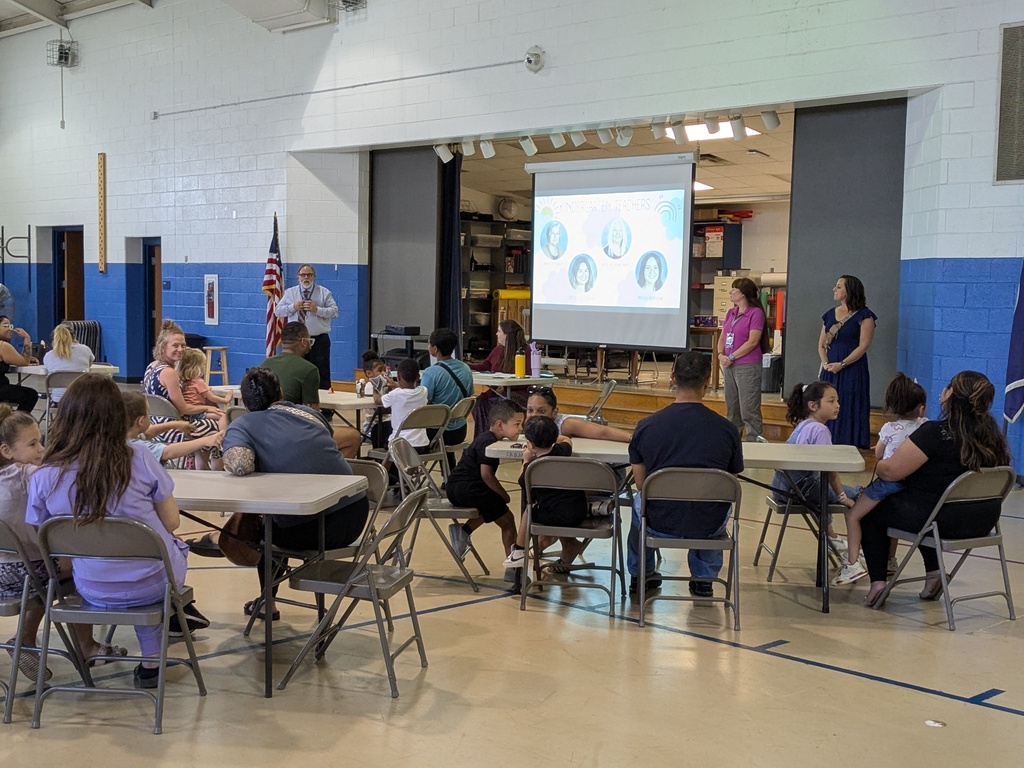 People sit in chairs around tables in a room, watching a presentation on a projector screen.