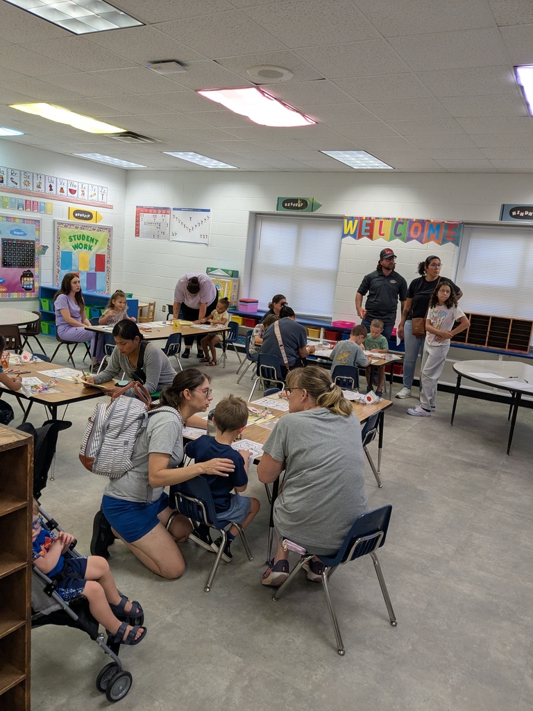 In a classroom, a group of children and adults are seated at tables. Some are seated in chairs and others are standing.