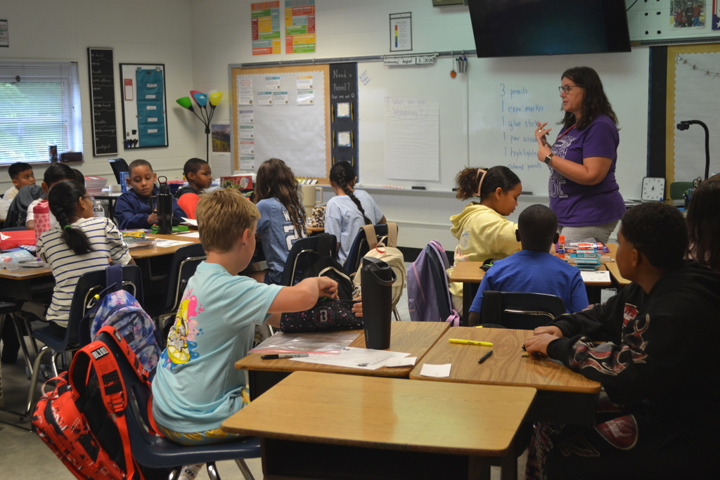 A classroom with a teacher addressing students at desks. Whiteboards and a mounted television are on the wall.