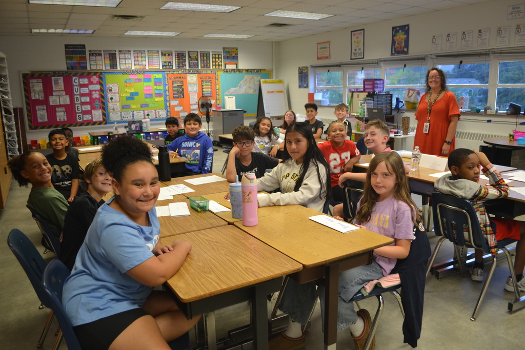 A classroom with students seated at desks, smiling. A teacher stands at the front, and walls are decorated with bulletin boards.