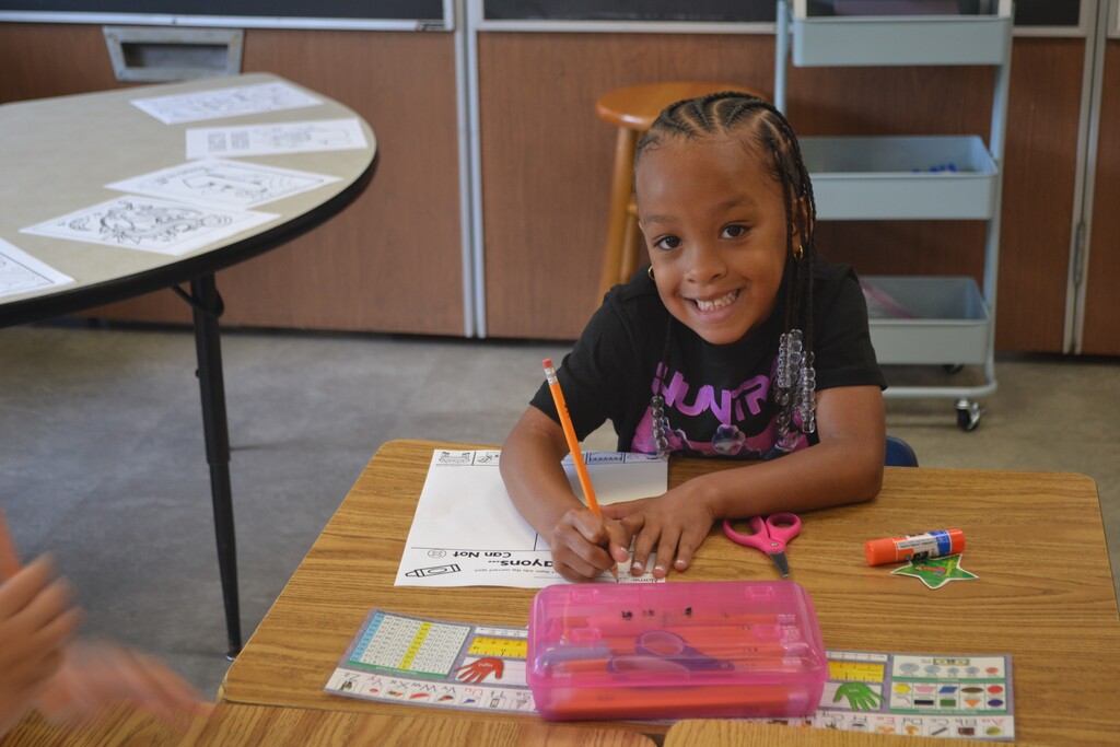 Student is smiling from her desk while holding a pencil writing on a worksheet.