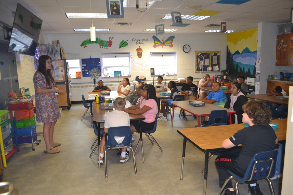 A classroom with children seated at tables. An adult stands at the front of the room.