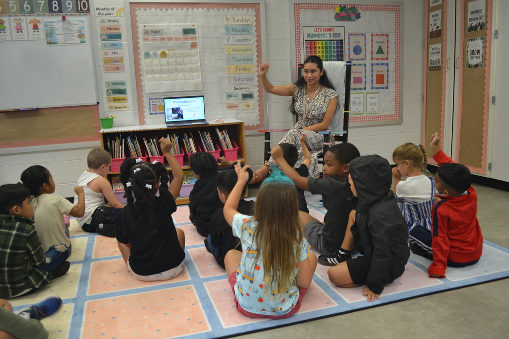 A woman sits on a chair in front of a classroom, with a laptop on a desk. Students sit on the floor, raising their hands to participate.