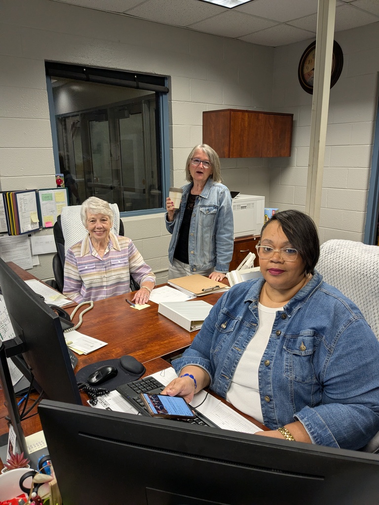 Three women at a desk in an office. One holds a cup, another holds a book, and the third sits with a computer.