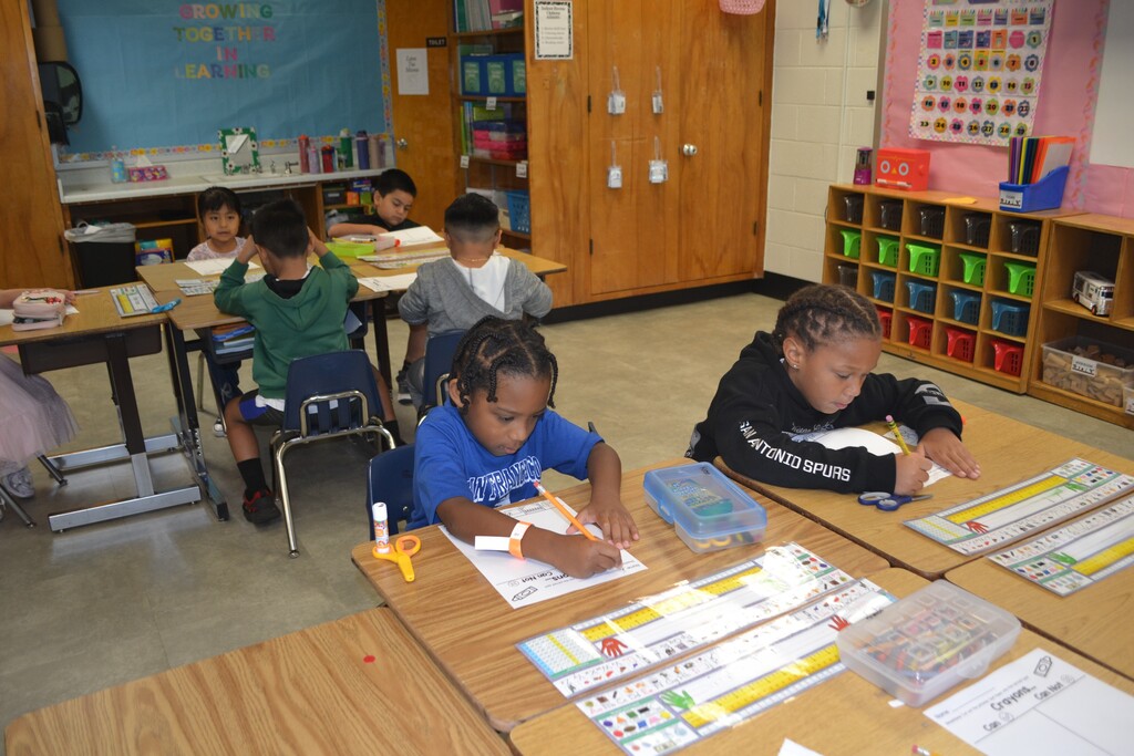 Children seated at desks in a classroom, engaged in activities. Rows of wooden cabinets and shelves with various items.