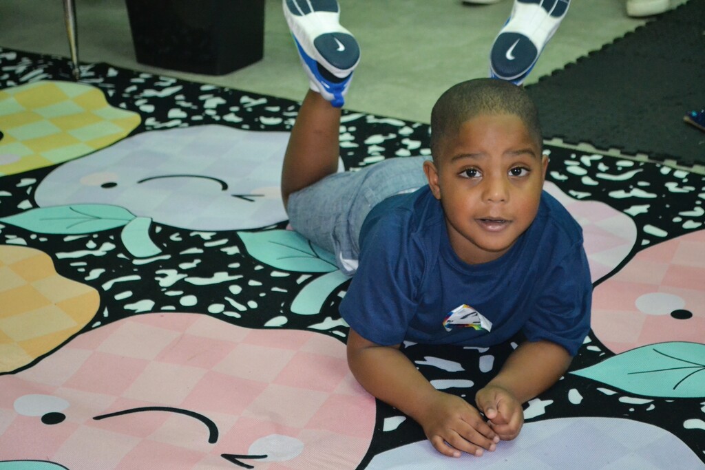 A child lies on a floor with a colorful rug. The rug has fruit and leaf designs. The child has sneakers on their head.