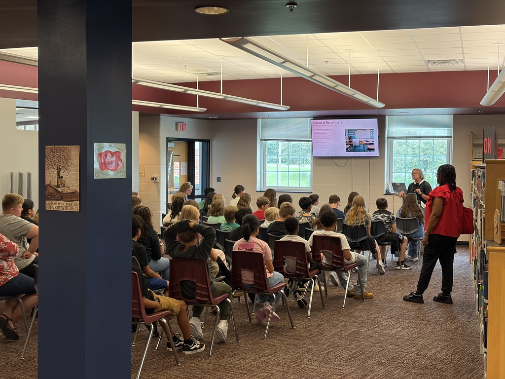 A group of students are seated in a circle in a room with a presentation screen. A woman stands nearby.