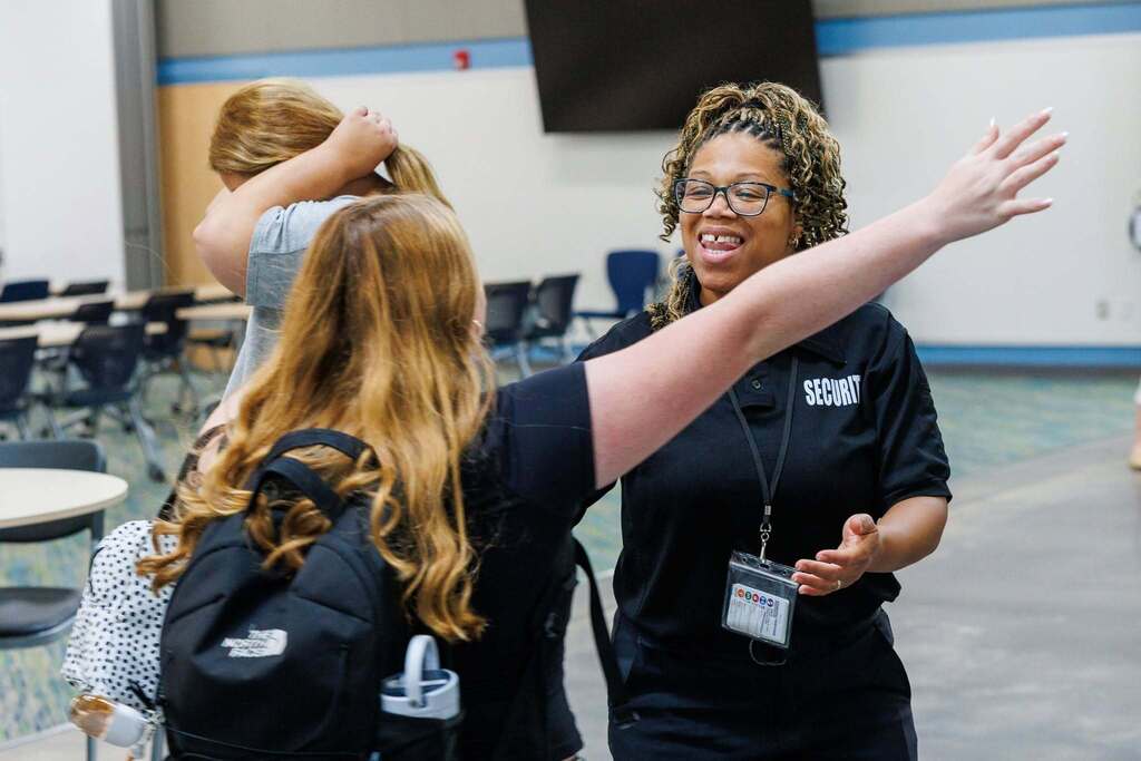 A security staff member warmly greets arriving students with open arms inside the Culpeper Technical Education Center, creating a welcoming atmosphere as students wearing backpacks gather at the start of the school day.