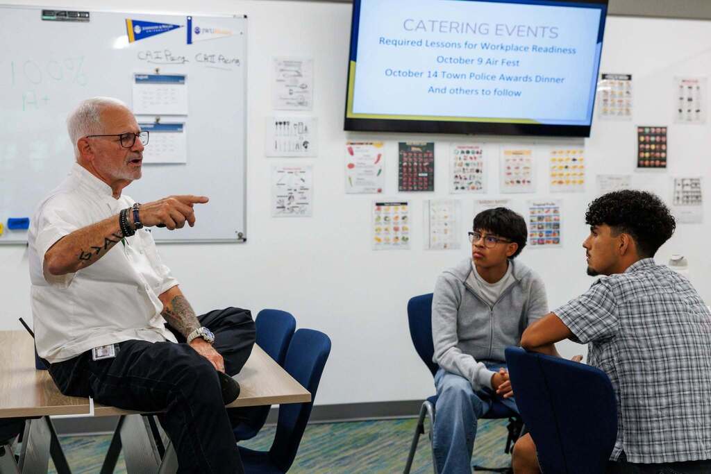 A Culinary Arts instructor speaks with two students in a classroom, leading a discussion about workplace readiness and upcoming catering events while students listen attentively.
