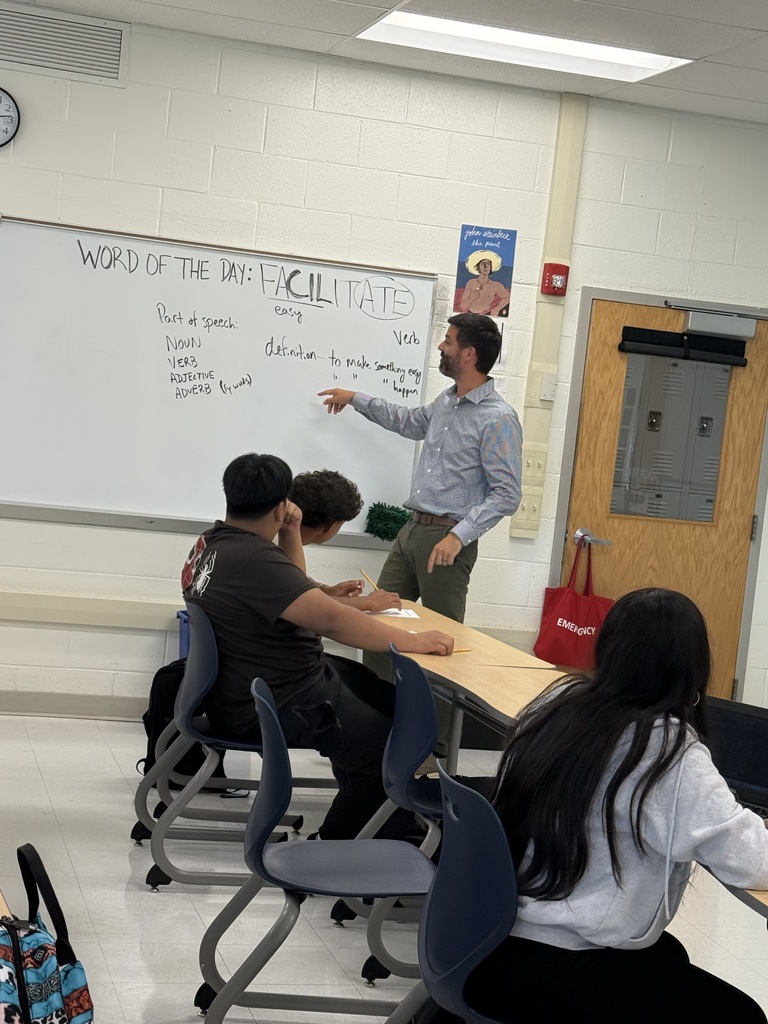 A classroom with a white board, a man teaching, and three students seated in chairs.