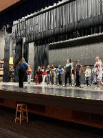 People gather on a stage in a theater, with a wooden stool on the left. Black curtains are in the background.
