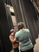 Two individuals face each other; one wears a hat, the other a teal shirt, with black theater curtains behind them.