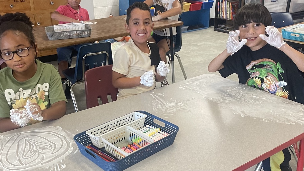 Three children with white gloves are seated at a table in a classroom. A table has drawings on it and a basket of art supplies.