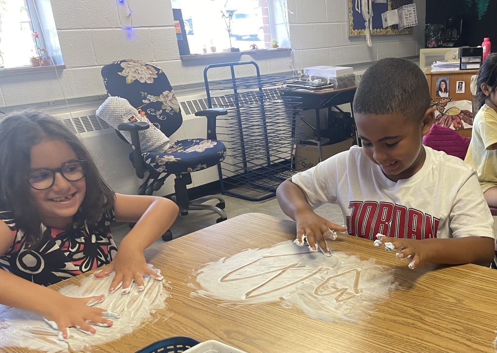 Two children sit at a table, one with glasses, both making art with flour in an indoor classroom.