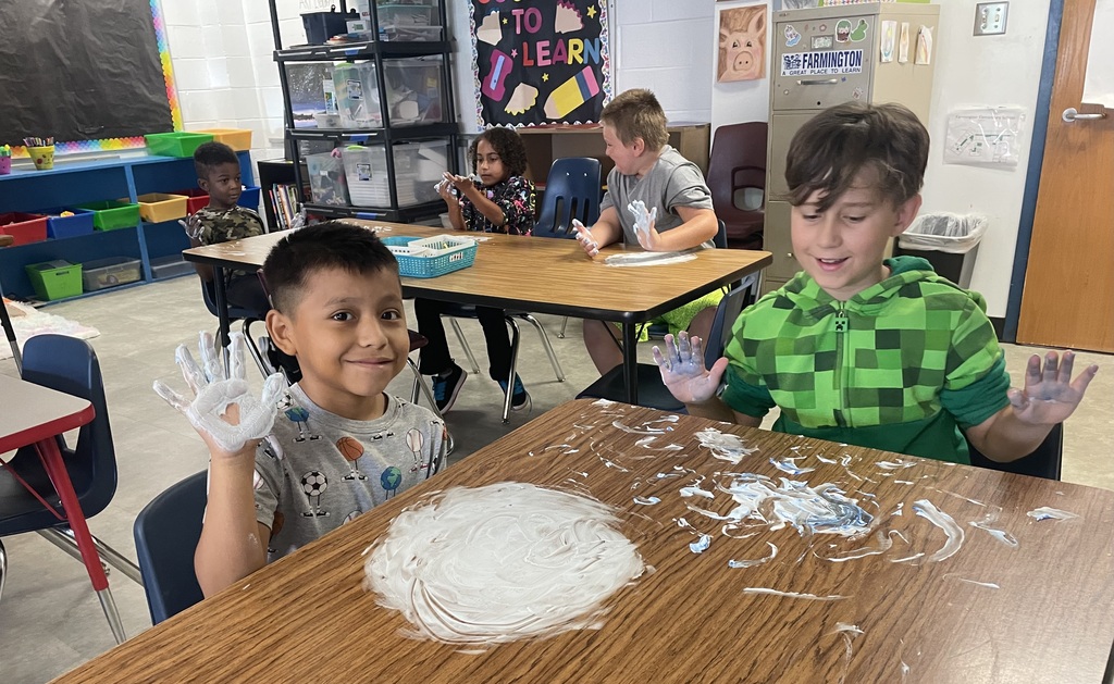 Two children sit at a table covered in paint. One child has paint on their hands. Other children sit at tables in a classroom.