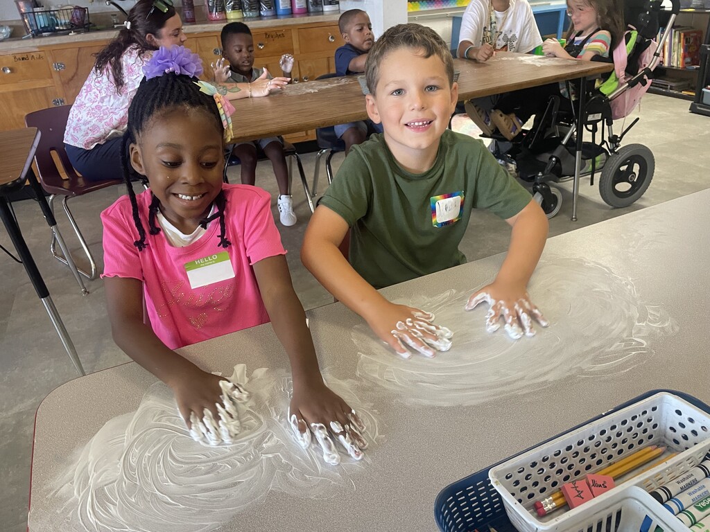 Two children sit at a table, playing with white substance, with several other children and adults around.