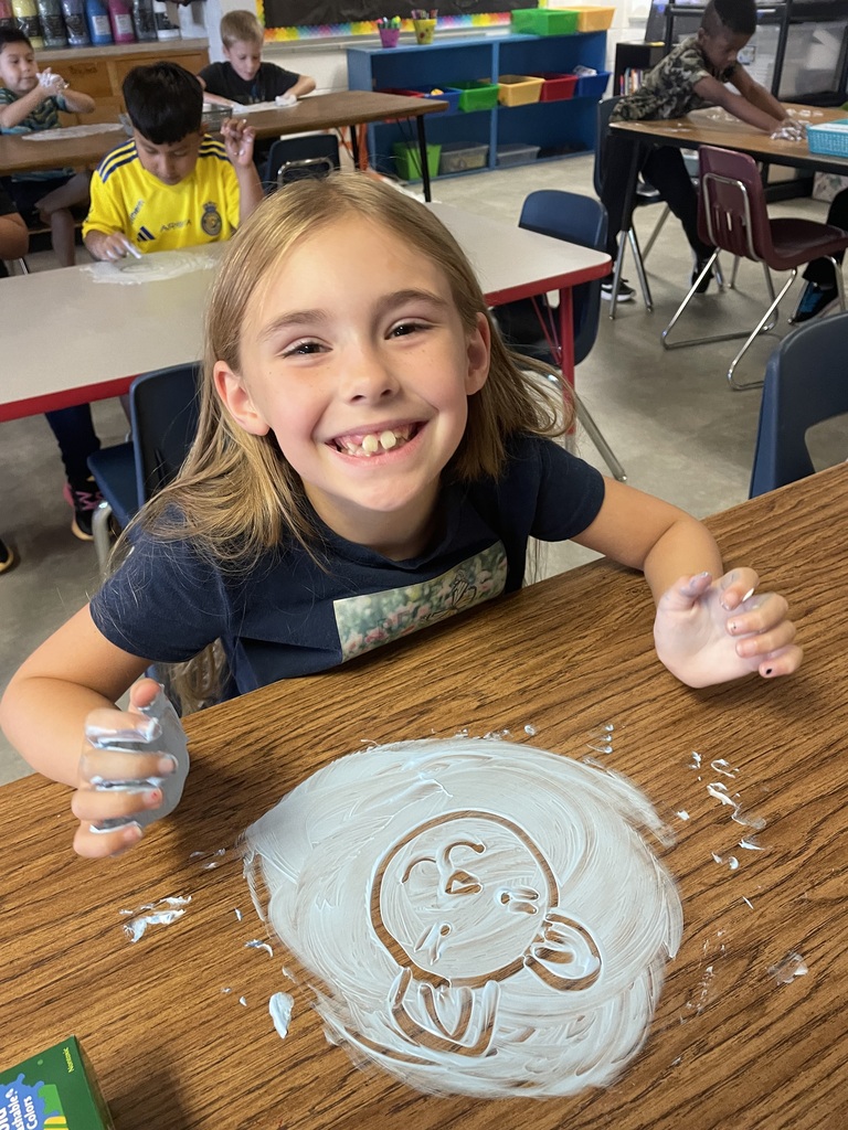 A young girl is sitting at a table, smiling, with a drawing of a turtle on paper.