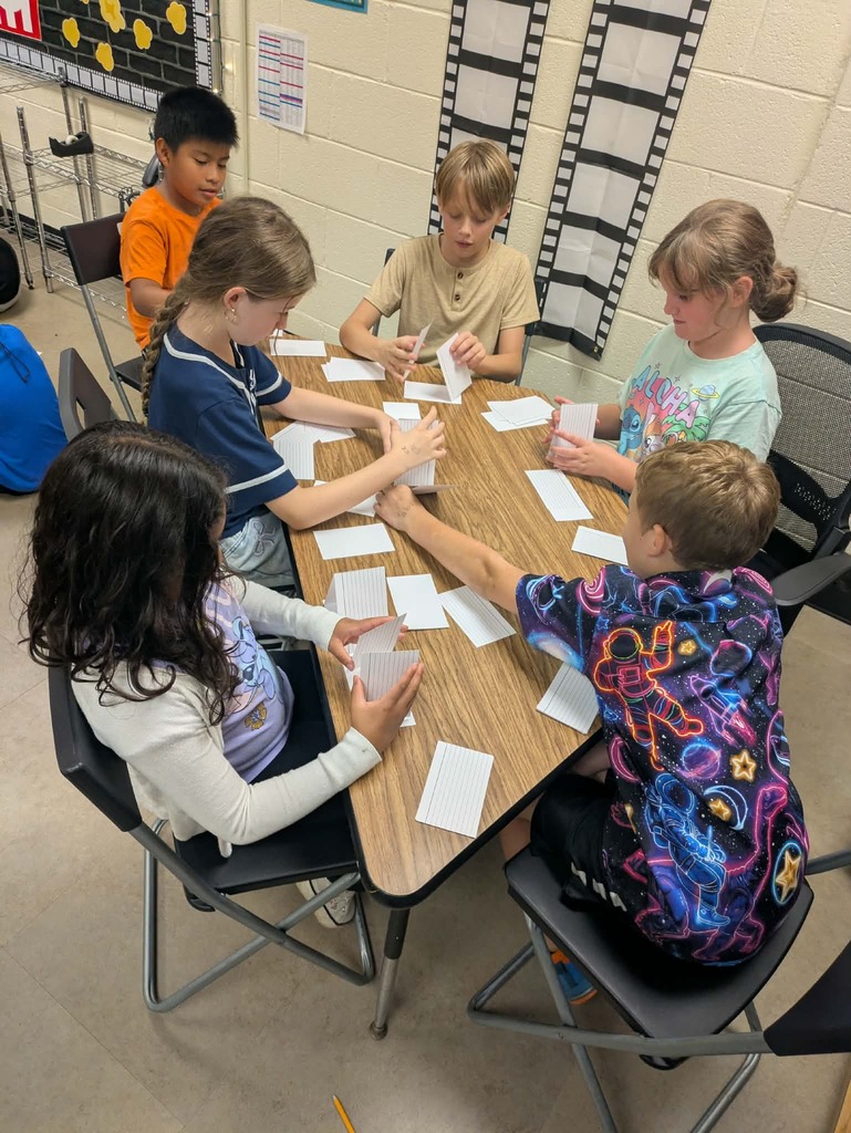 Six children seated around a table, holding papers and playing a game. One child is reaching for another's paper.