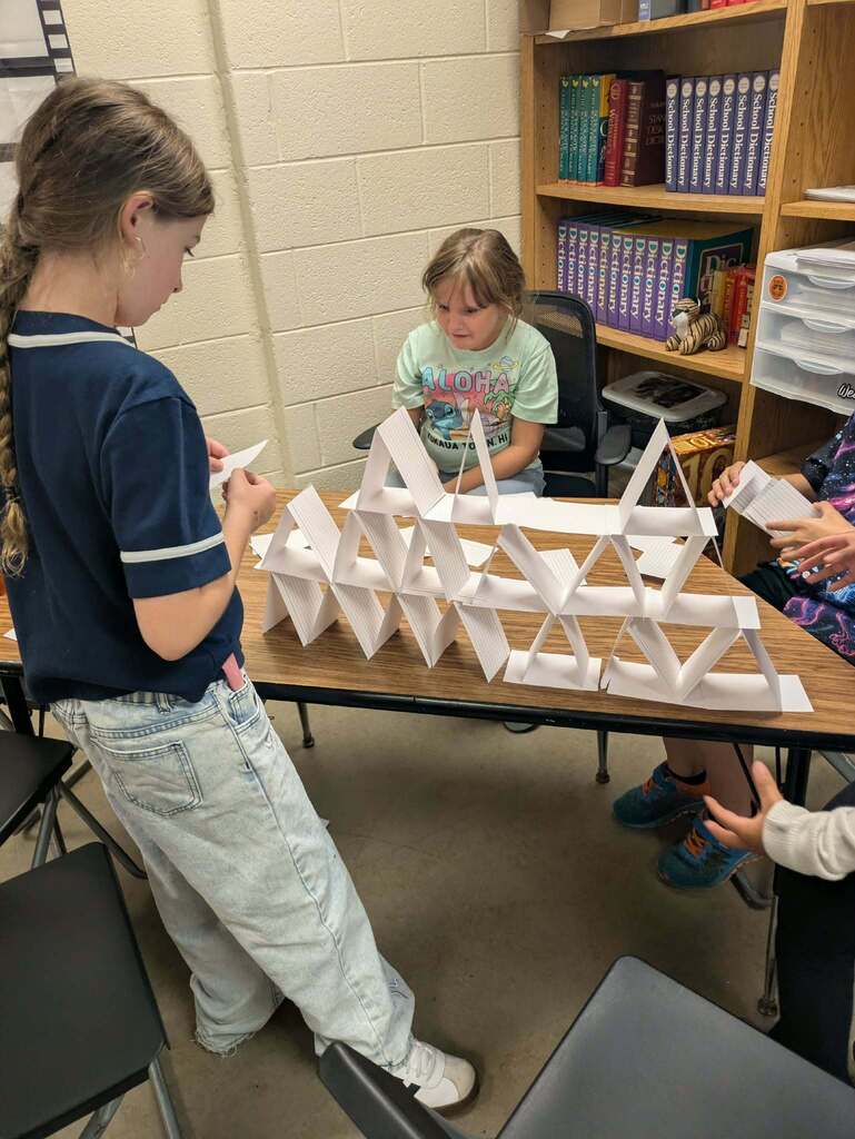 Three children work together at a table, constructing a structure with white paper. Behind them, shelves filled with books and a chair.