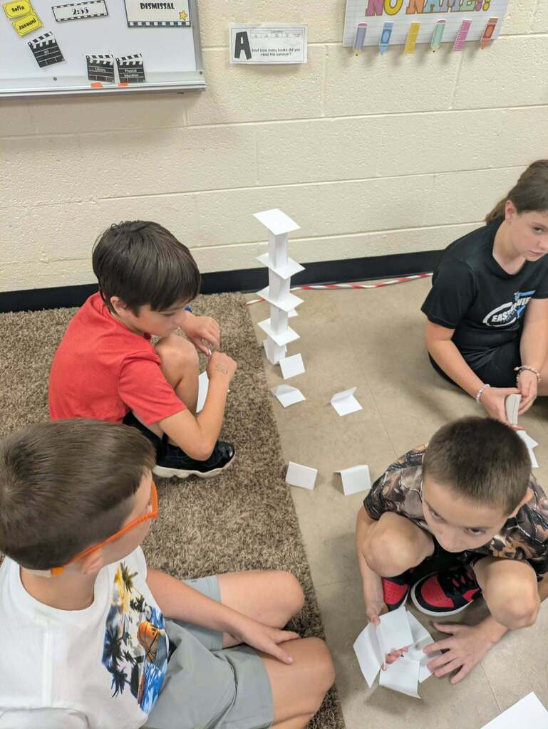 Four children are seated on the floor constructing a tower of cards. One boy wears glasses, another a bracelet.