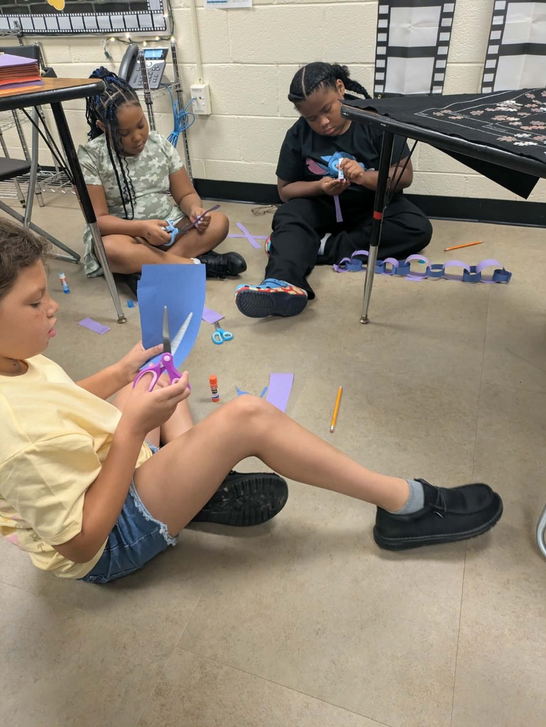 Children sit on the floor in a classroom. One uses scissors on paper, another holds a craft.