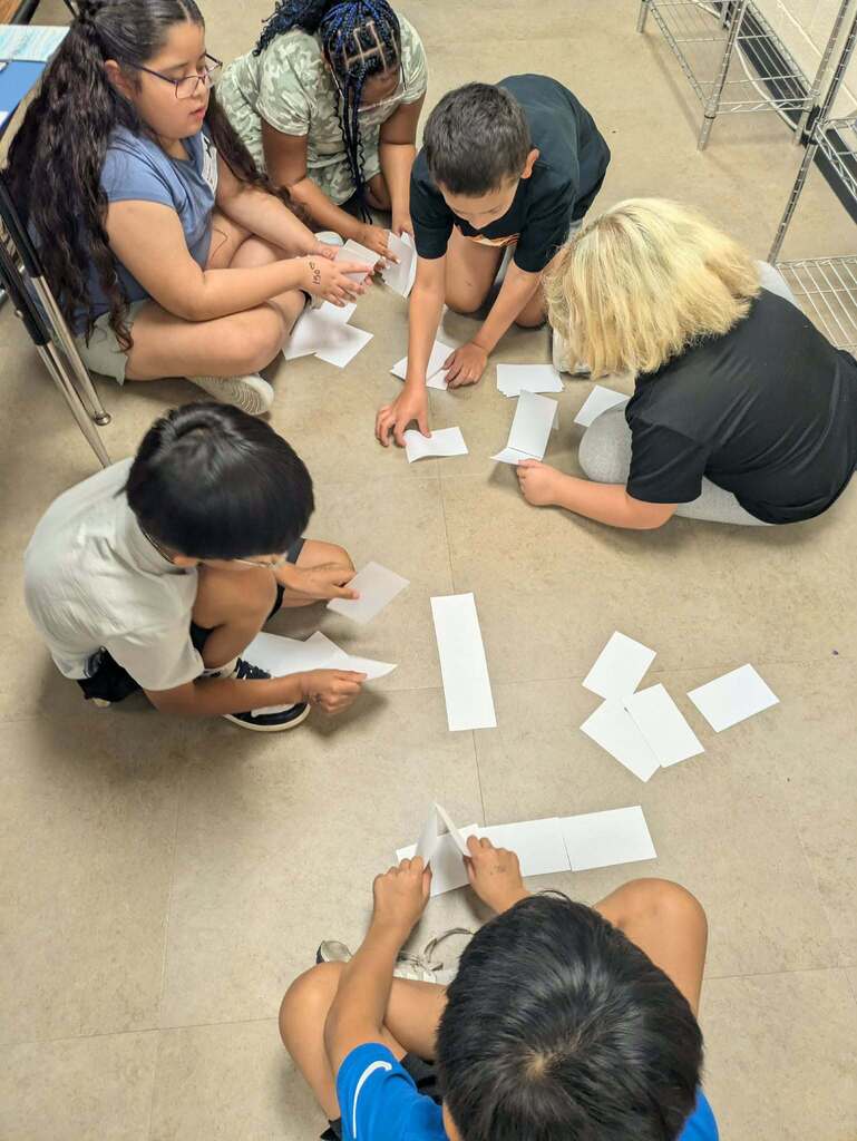 Five children and an adult sitting on a classroom floor with pieces of paper. One child holds a paper, and others gather around.