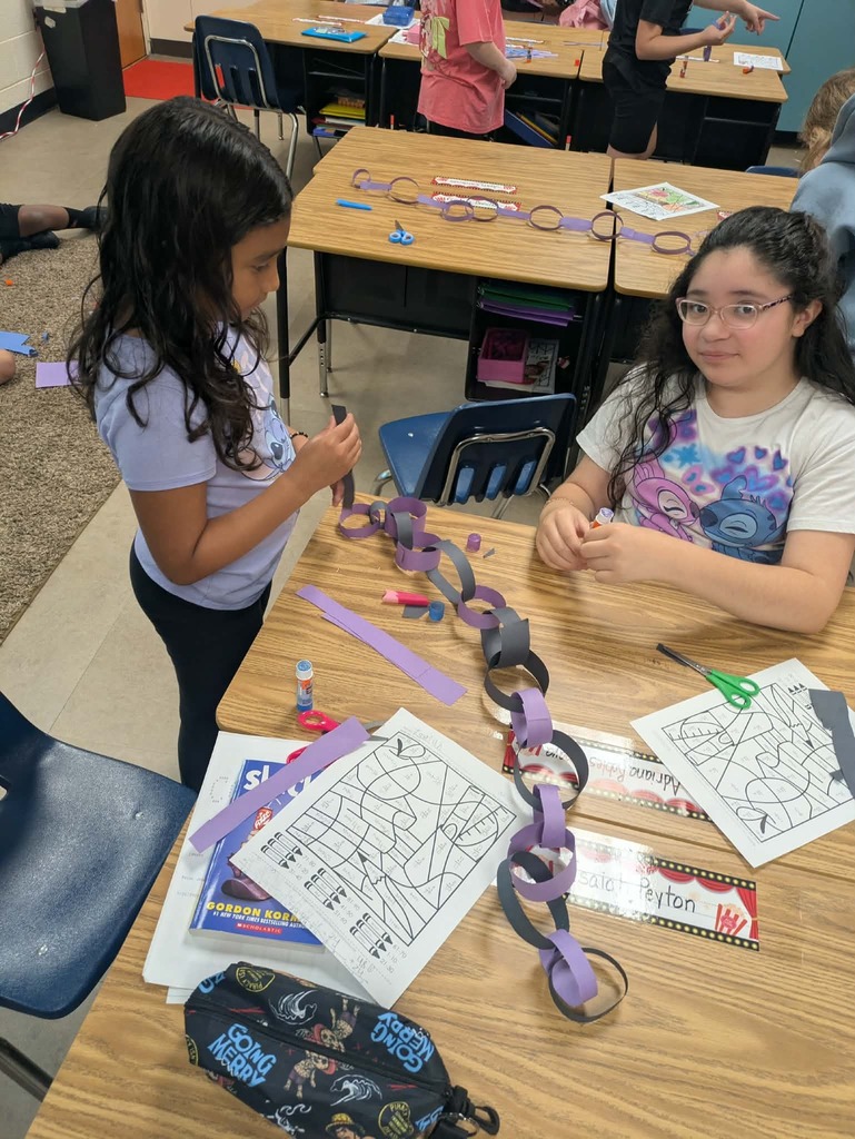 Two girls seated at a table, working on crafts. One girl has a mask on the table, and they both hold scissors.